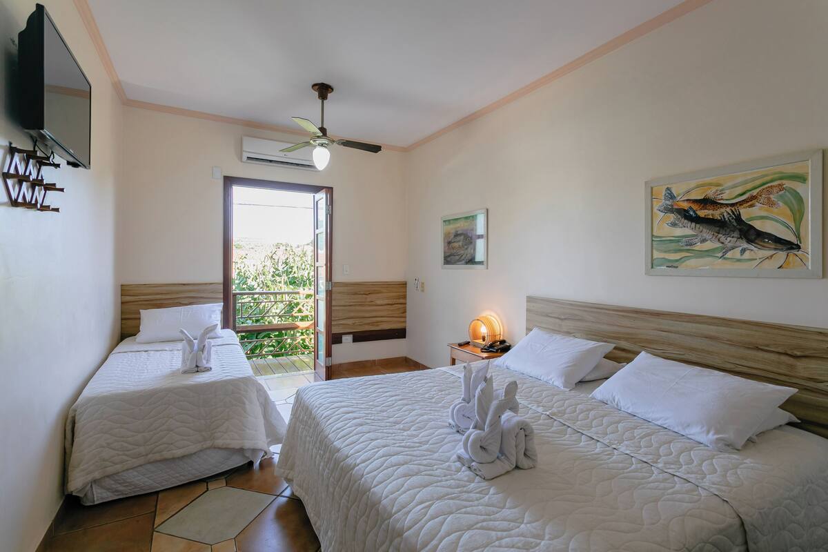 A hotel room with two beds, a ceiling fan, an air conditioning unit, a painting, and a view of greenery outside.