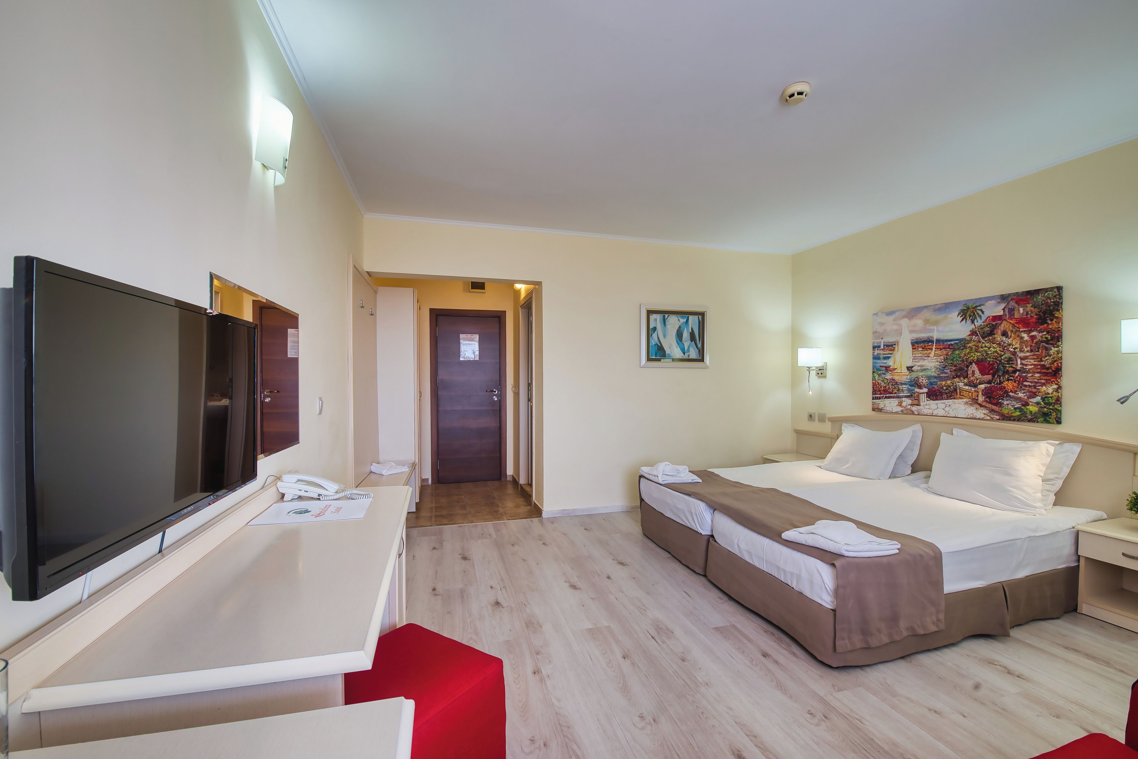 A hotel room with a bed, a desk, a TV, a painting, and a door leading to another room.