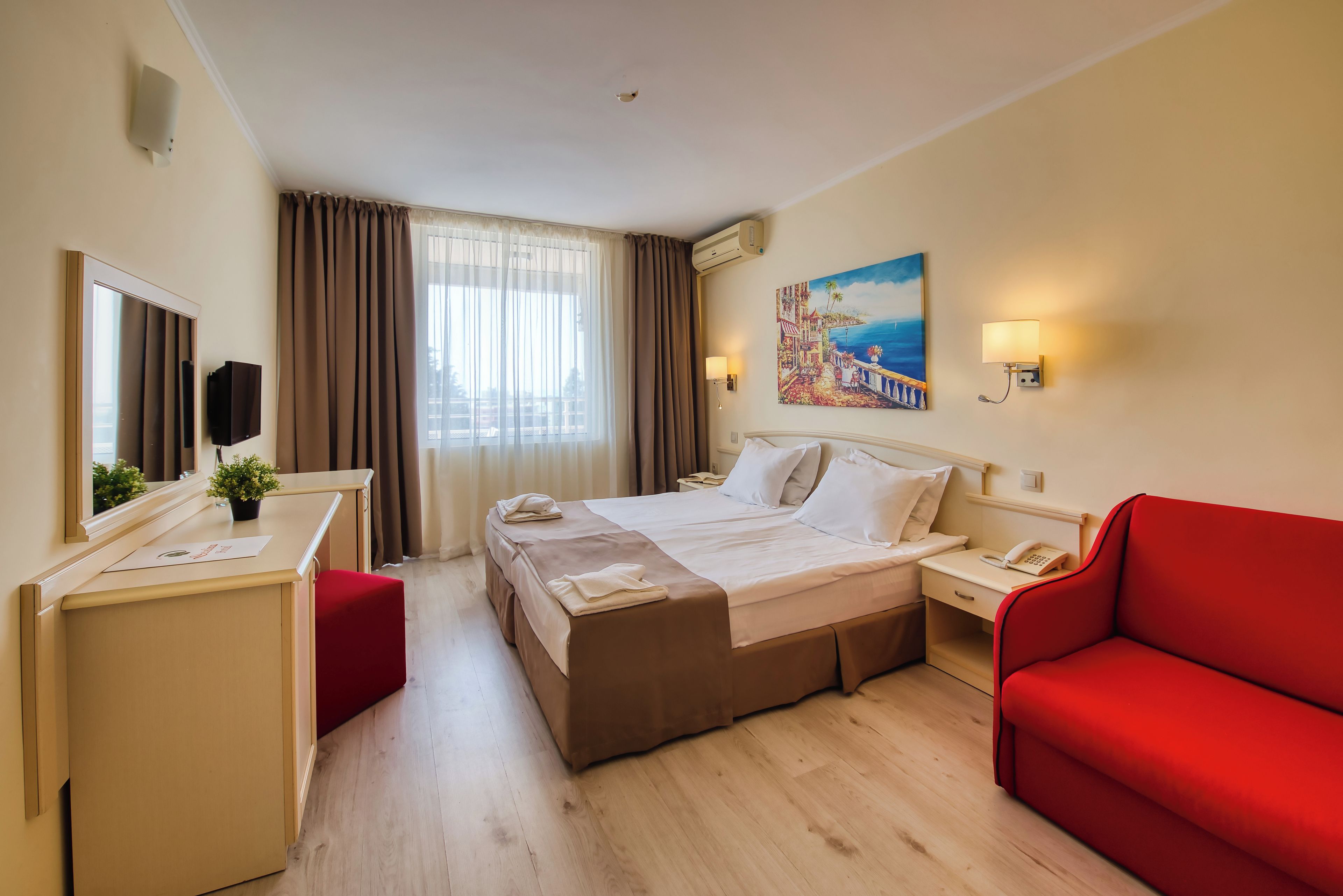 A hotel room with a bed, a desk, a red sofa, a painting, and a window with curtains.