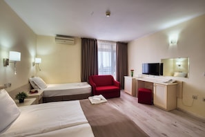 A hotel room with two beds, a desk with a TV, a red chair, and a small red ottoman. - Detelina (Varna)