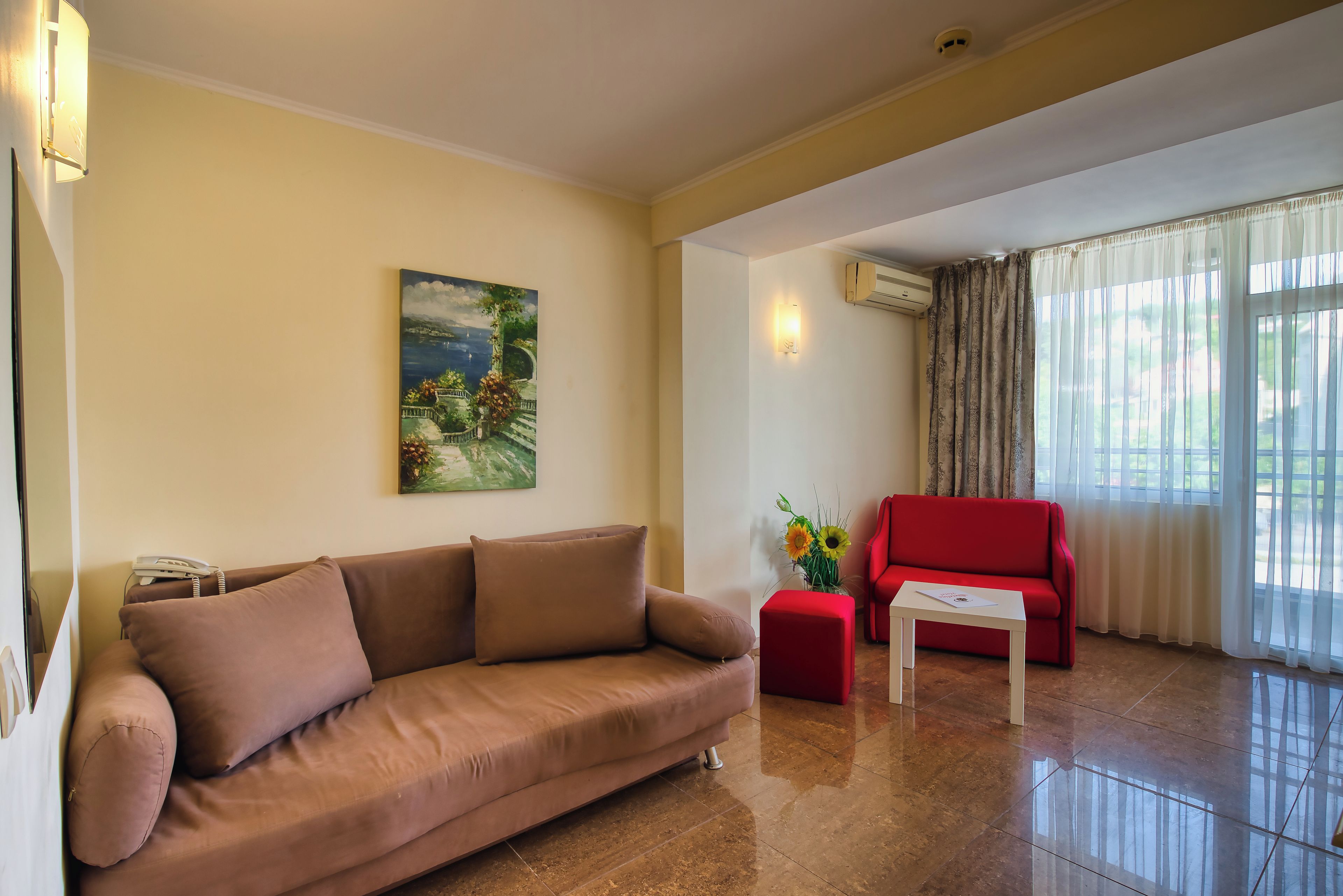 A living room with a sofa, a red armchair, a coffee table, a painting on the wall, and an air conditioning unit.