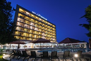 Un hotel con múltiples balcones, una zona de piscina con tumbonas y sombrillas, y un exterior bien iluminado.