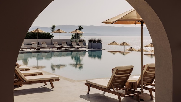 Piscina infinita con sillones de descanso y sombrillas, con vista despejada al mar y montañas distantes.