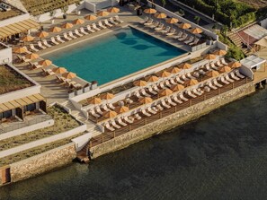An aerial view of a resort with a large swimming pool surrounded by sun loungers and umbrellas, a stone wall along the water's edge, and a covered seating area with tables and chairs.
