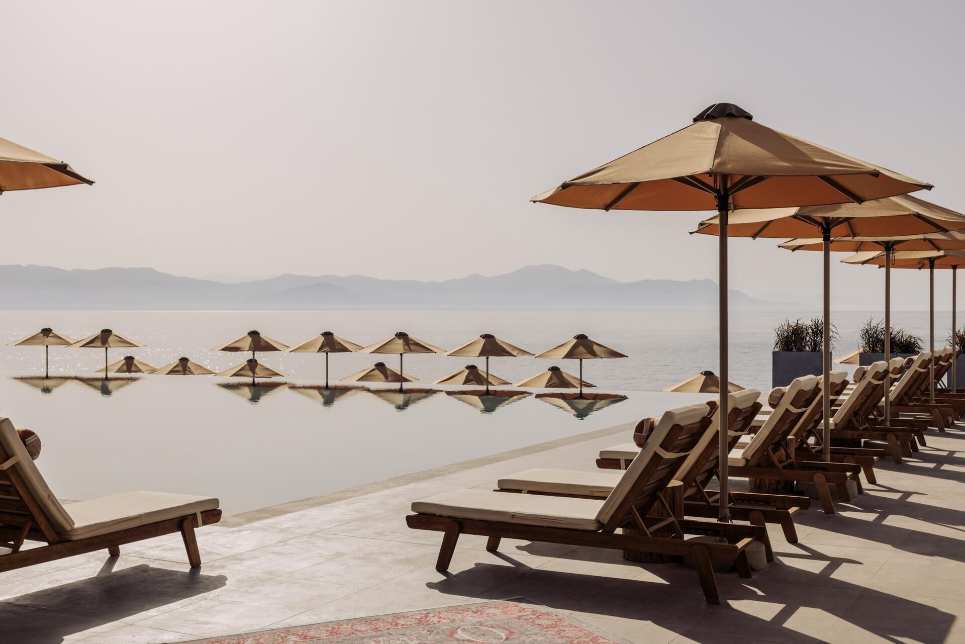 Piscina infinita con sillones de descanso y sombrillas, con vista a una masa de agua tranquila y montañas lejanas.