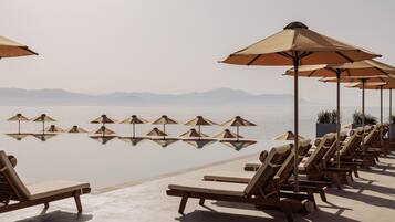 Piscina infinita con sillones de descanso y sombrillas, con vista a una masa de agua tranquila y montañas lejanas.