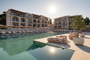 Ein Poolbereich mit Liegestühlen und Sonnenschirmen, ein Gebäude mit mehreren Balkonen und Kübelpflanzen.