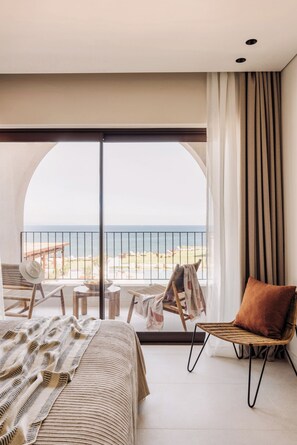 Ein Hotelzimmer mit großem Fenster, einem Bett, einem Sessel, einem kleinen Tisch und Blick auf den Strand.