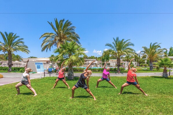 A group of people performing yoga in an outdoor grassy area with palm trees and buildings in the background. - Kipriotis Maris Suites by Anayia All Inclusive Resorts (Kos)