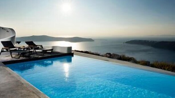 A swimming pool with lounge chairs and a view of the sea and distant land. - Avaton Resort and Spa (Santorini)