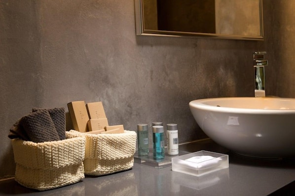 A bathroom with a large white sink, a mirror, and a textured wall. There are two woven baskets with toiletries and a clear plastic container on the counter. - Avaton Resort and Spa (Santorini)