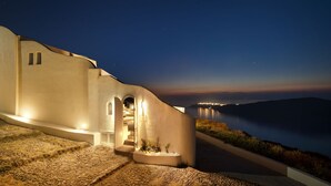 A coastal building with a staircase leading to a lit entrance, overlooking a calm sea and distant city lights. - Avaton Resort and Spa (Santorini)