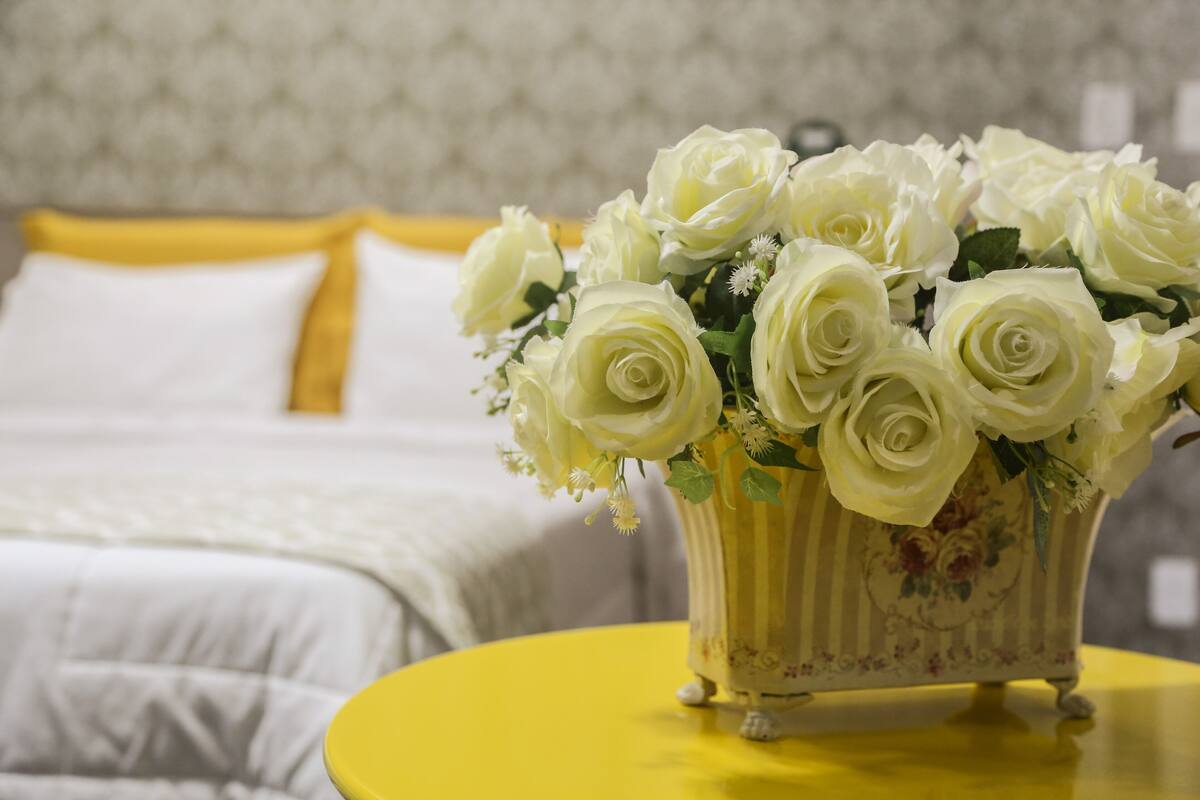 Una cama con ropa de cama blanca y un almohadón amarillo, una mesa amarilla con un florero de rosas blancas, y una pared con un diseño llamativo al fondo.