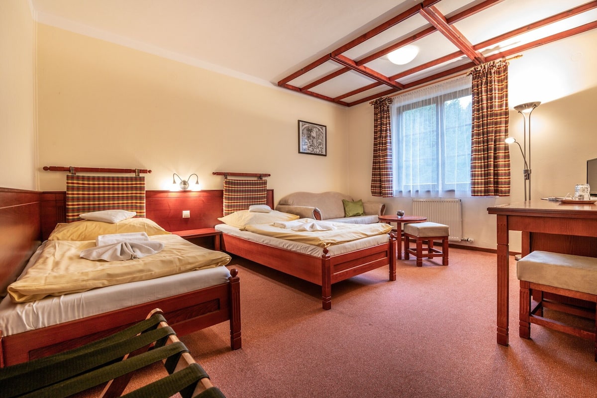 A hotel room with two wooden beds, a small table, a chair, a lamp, and a window with curtains.