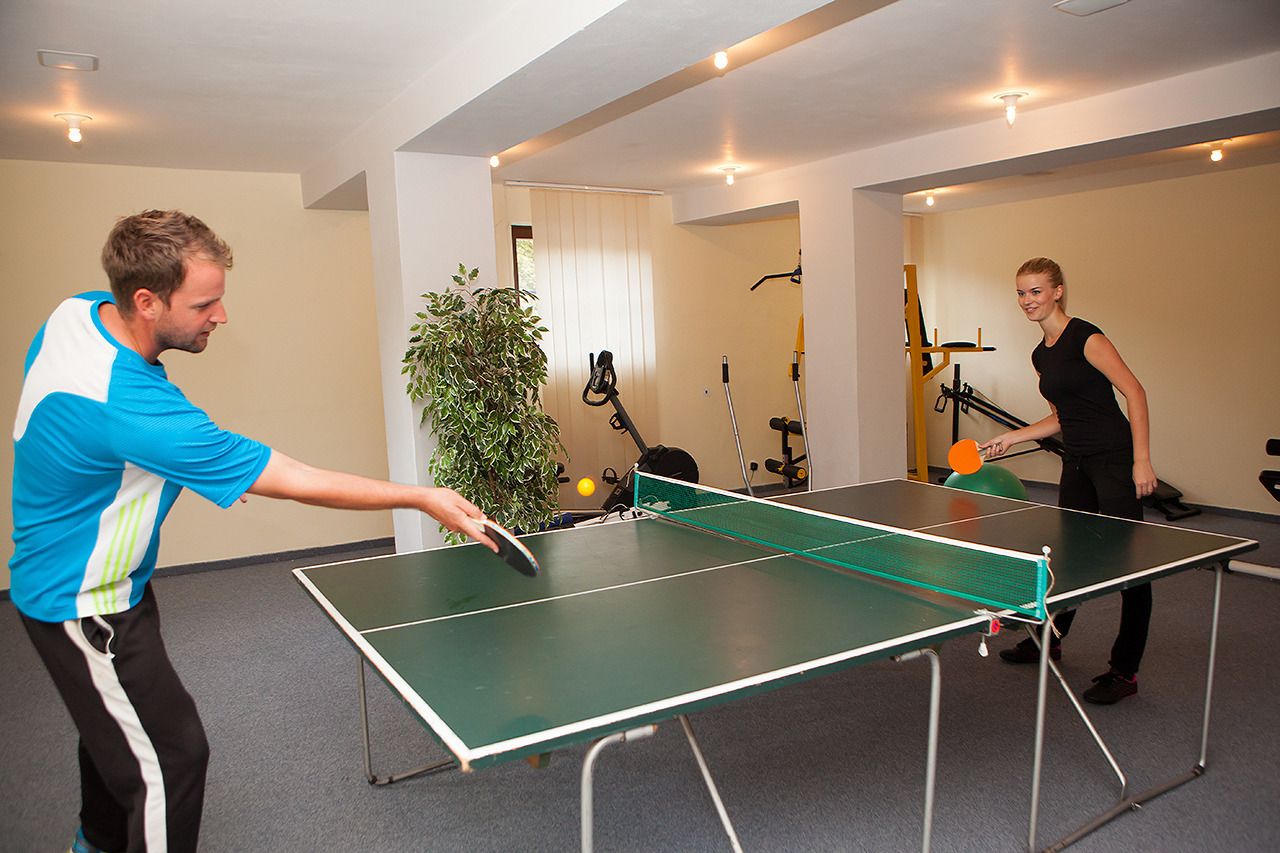 Twee personen spelen tafeltennis in een ruimte met een tafeltennistafel, trainingsapparatuur en een potplant.