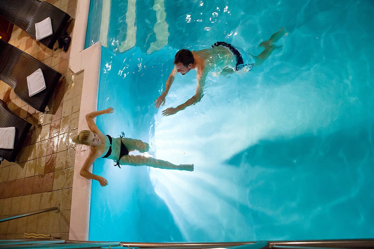 A swimming pool with two people swimming, lounge chairs, and a tiled floor.