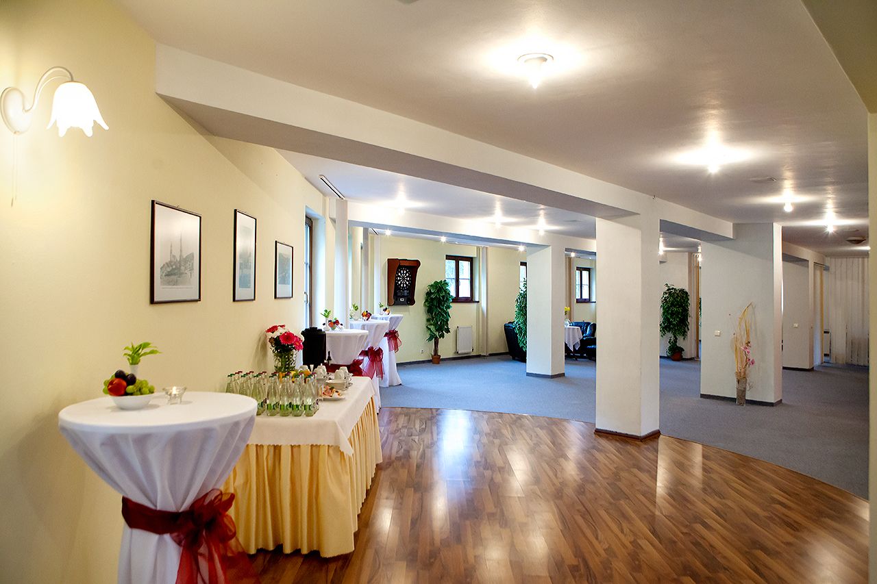 A spacious hall with wooden floors, white walls, and framed pictures. Tables are set up with floral arrangements and a fruit bowl.