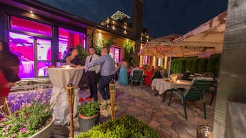 An outdoor patio with tables and chairs, surrounded by potted plants and a brick floor.