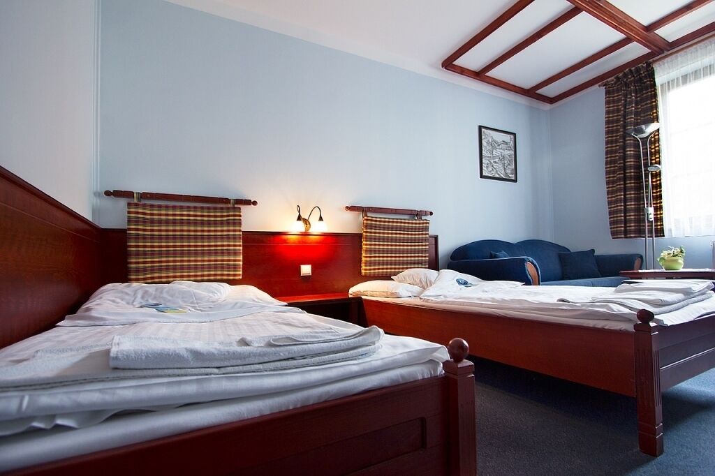 A hotel room with two wooden beds, a nightstand with a lamp, a blue sofa, and a window with curtains.