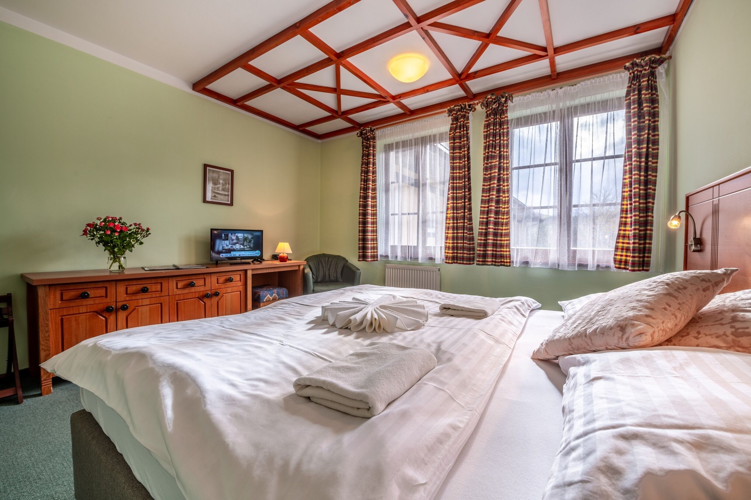 A well-lit bedroom with a wooden ceiling, a bed with neatly arranged towels, a desk with a television, and a window with patterned curtains.