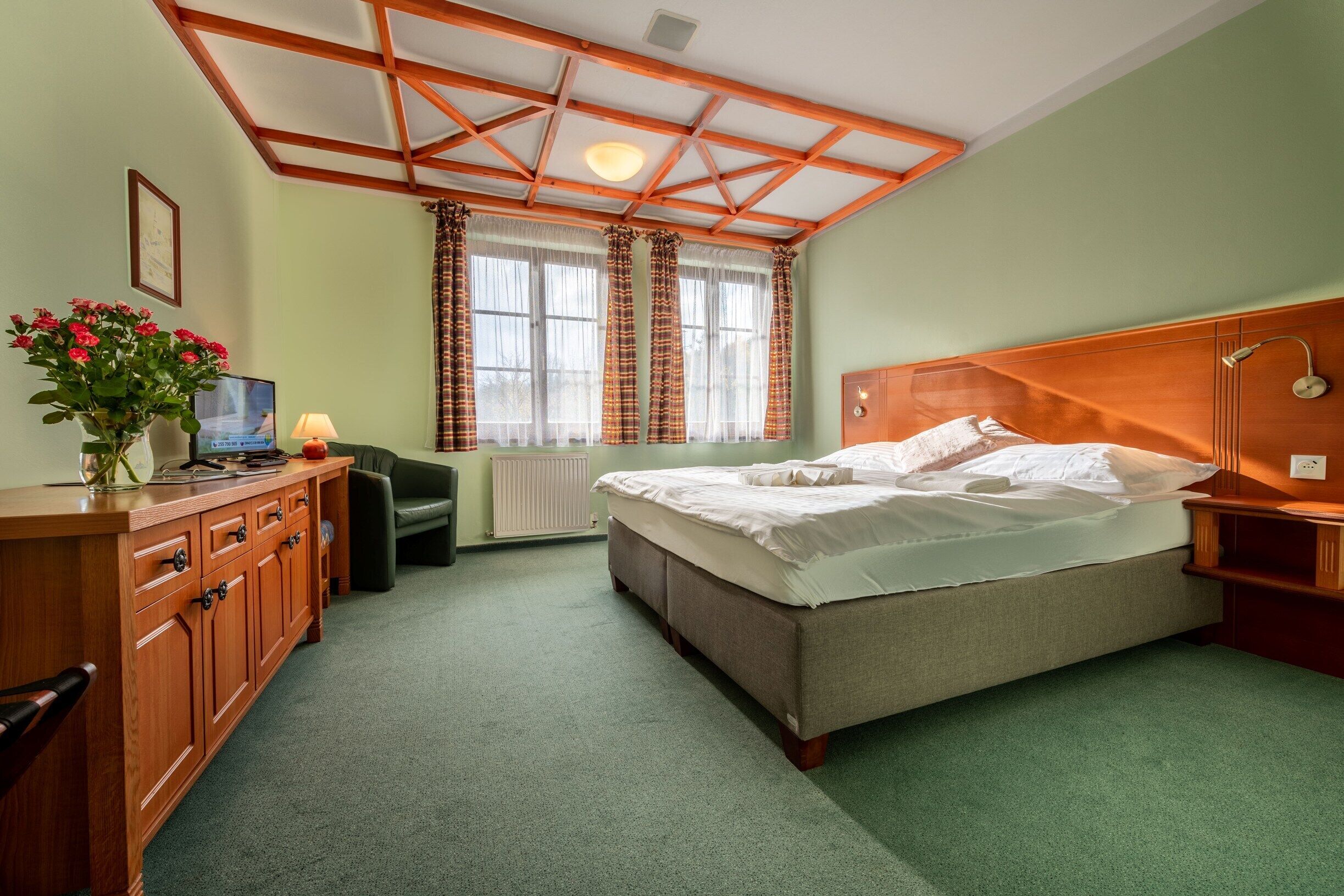 Une chambre d’hôtel avec une tête de lit en bois, un lit avec du linge de maison blanc, une commode en bois, un bureau avec un ordinateur, une chaise, un radiateur et une fenêtre avec des rideaux.
