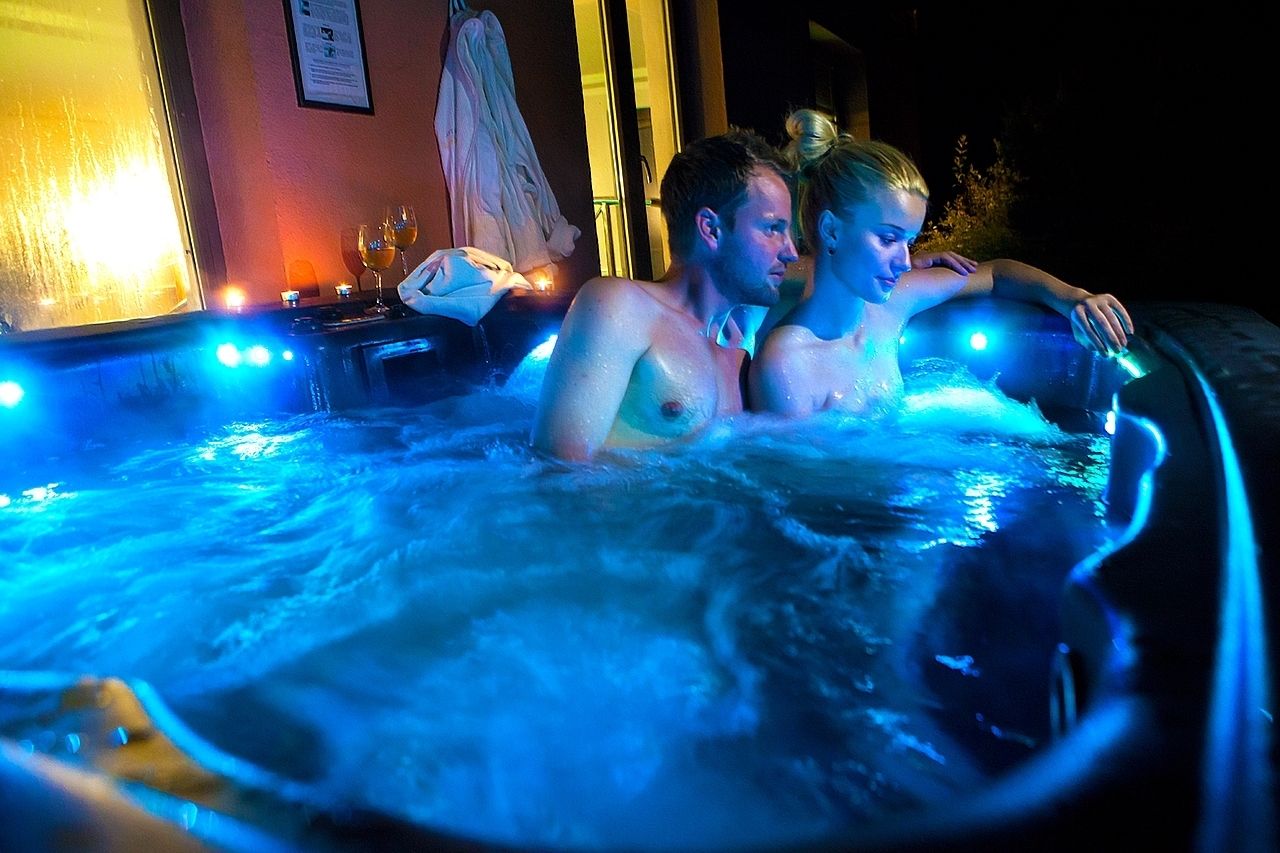 A couple in a hot tub with blue lighting, a towel, and a glass of wine on the side.