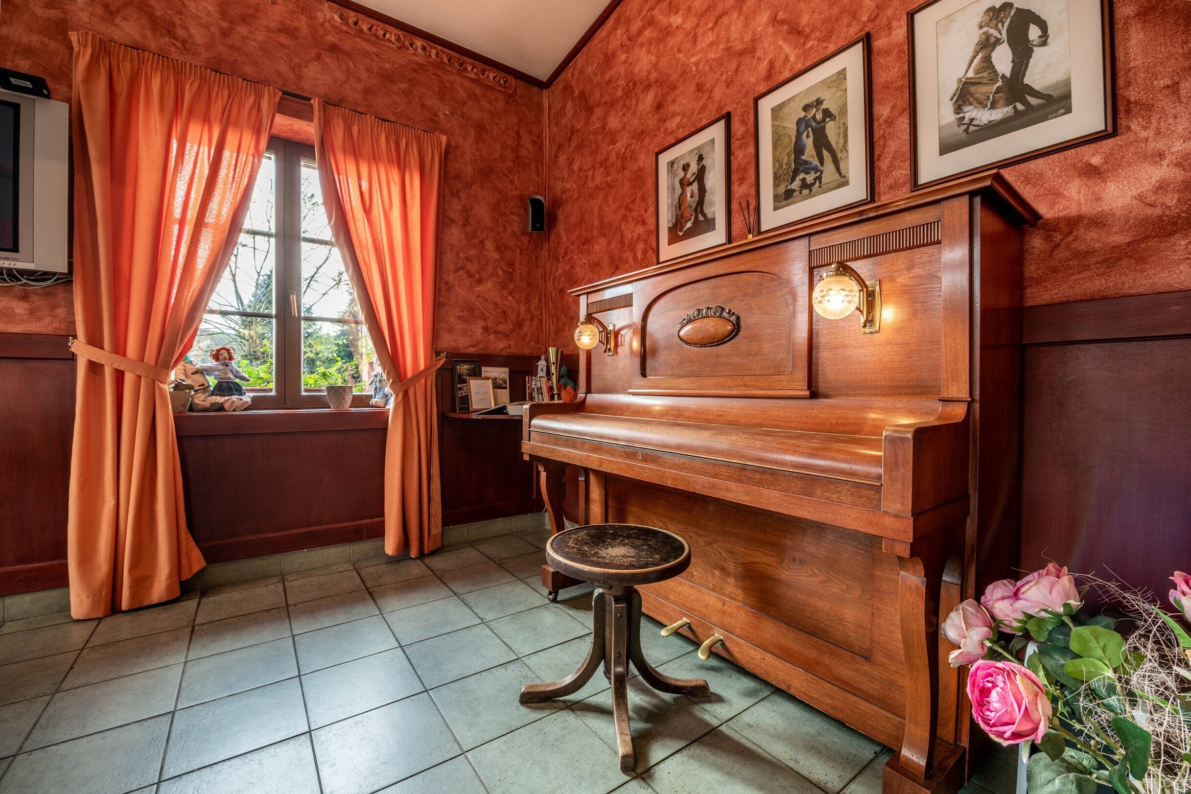 A room with a wooden piano, a stool, a window with curtains, and framed pictures on the wall.