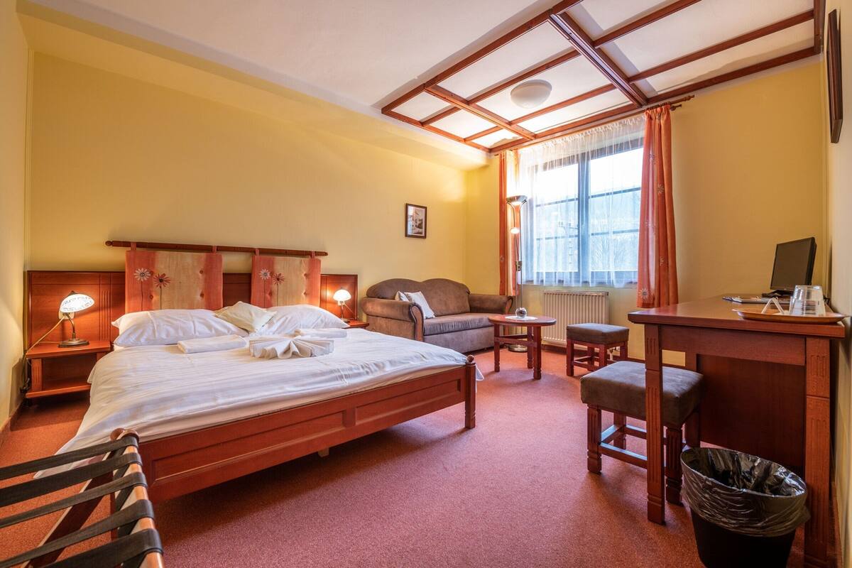 A hotel room with a bed, a desk, a chair, a small table, a sofa, and a trash bin.
