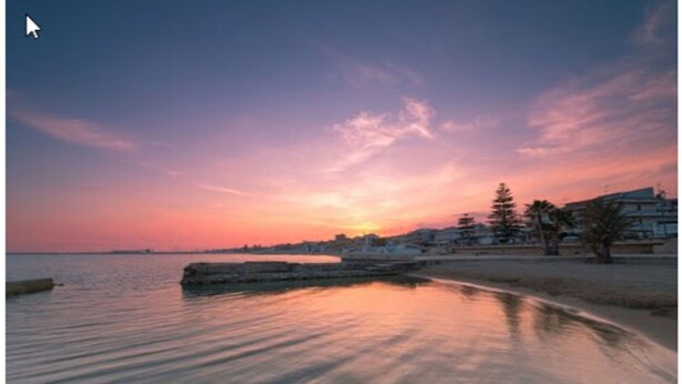 Un coucher de soleil sur la côte, avec des eaux calmes reflétant le ciel, une promenade et des immeubles d’habitation.
