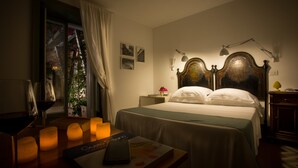 Une chambre d’hôtel avec un lit, des lampes de chevet, une table de nuit, une fenêtre avec des rideaux, et une table avec des bougies et un livre.
