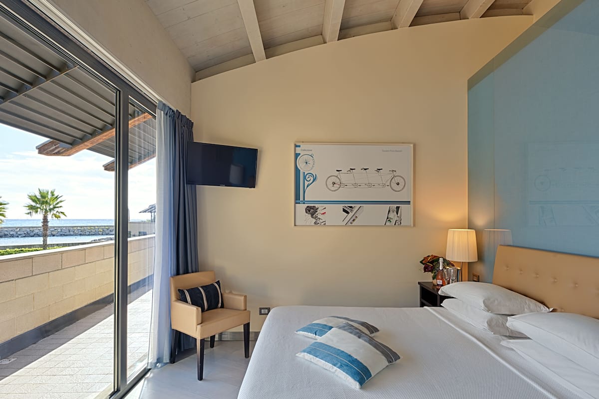 A hotel room with a large bed, a chair, a TV mounted on the wall, and a view of the ocean through a sliding door.