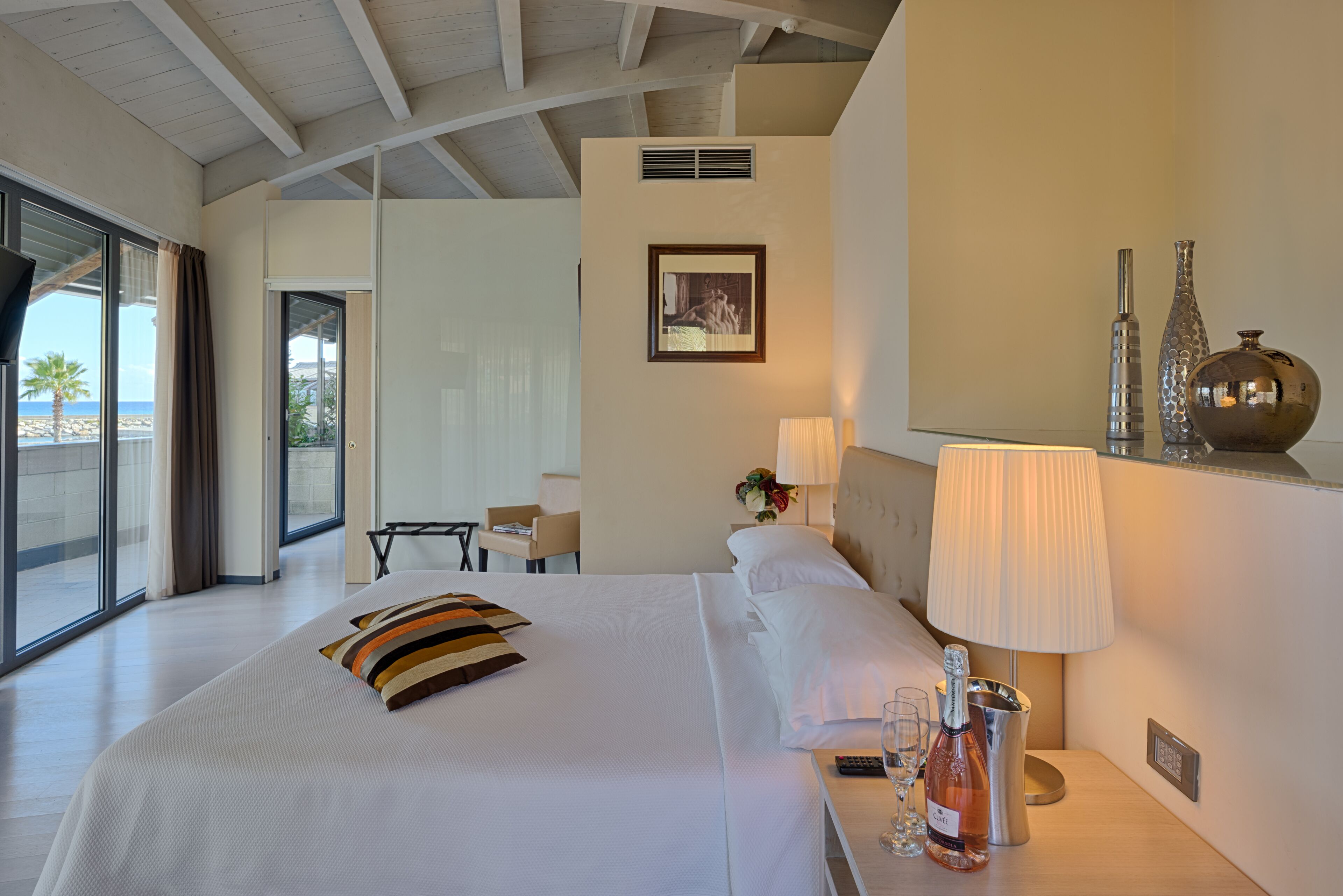 Une chambre d’hôtel avec un grand lit, une table de chevet avec une bouteille et des verres, une lampe, et une vue sur la plage par une grande fenêtre.