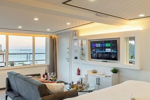 Une chambre d’hôtel dotée d’une grande télévision diffusant divers services de streaming, d’un canapé, d’une table à manger avec une bouteille et des verres, et offrant une vue sur la mer grâce à de grandes fenêtres.