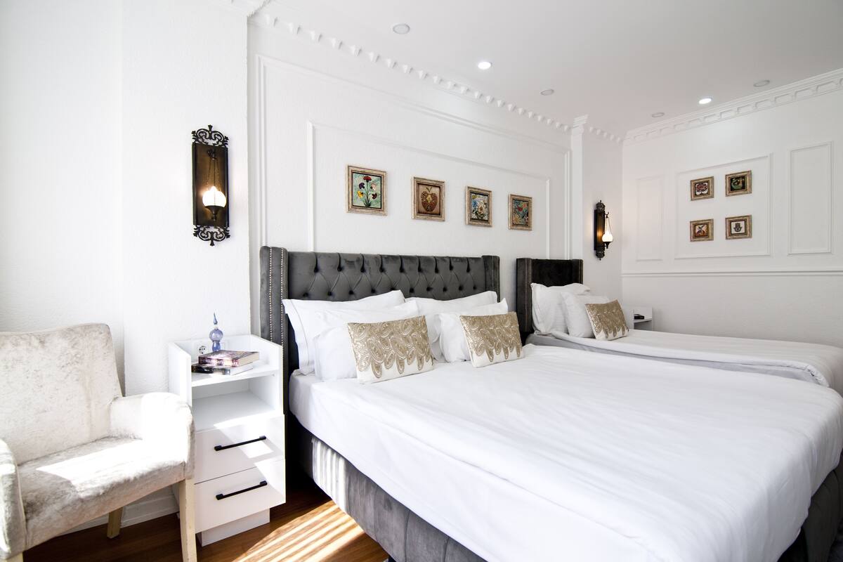 A hotel room with two beds, a white sofa, a bedside table with a lamp, and framed artwork on the walls.