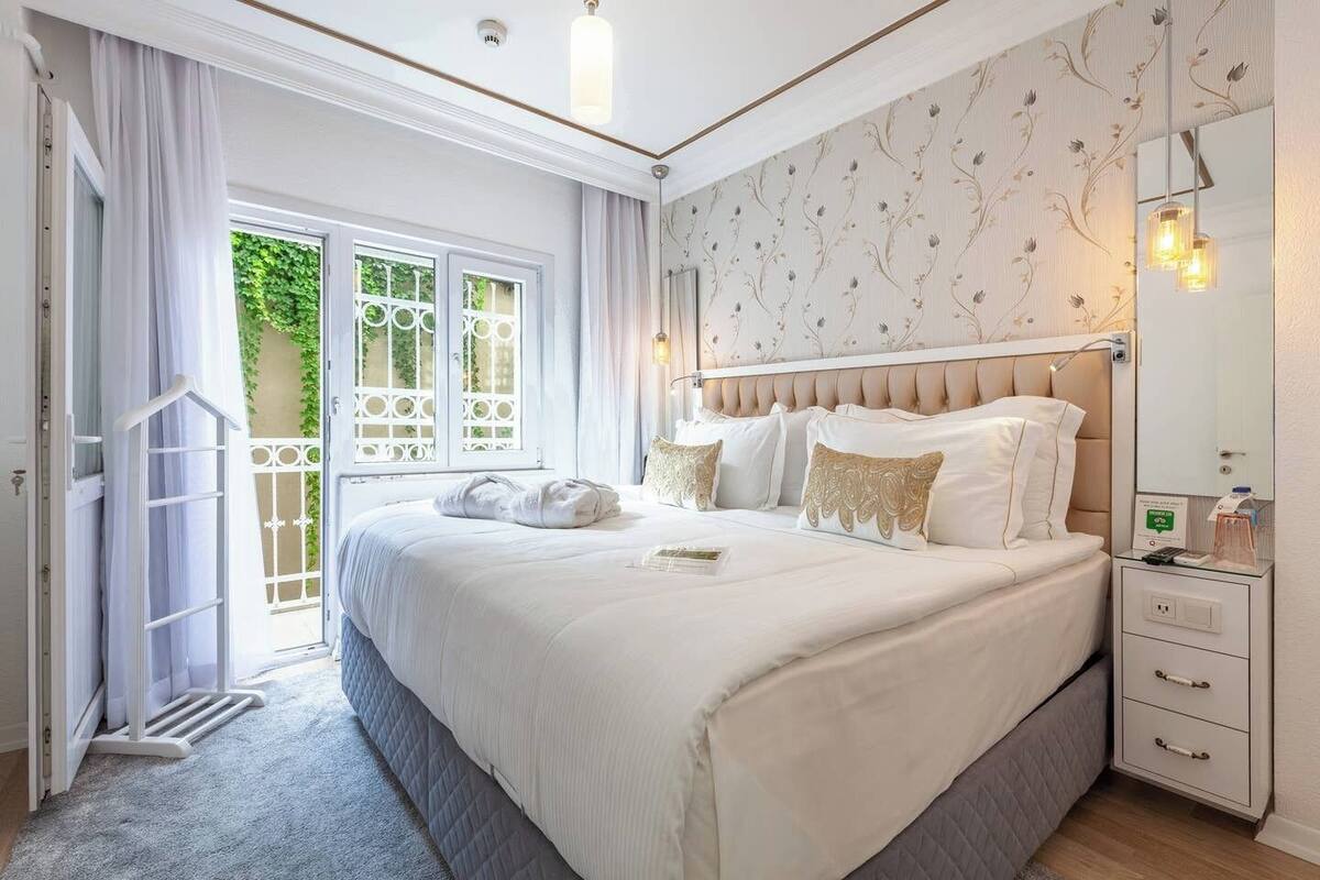 A spacious bedroom with a large bed, a bedside table with a mirror, and a view of greenery outside.