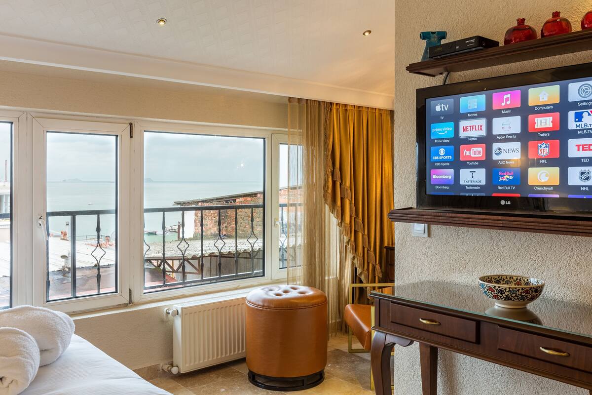 A hotel room with a large window, a television, a small stool, and a decorative bowl on a dresser.