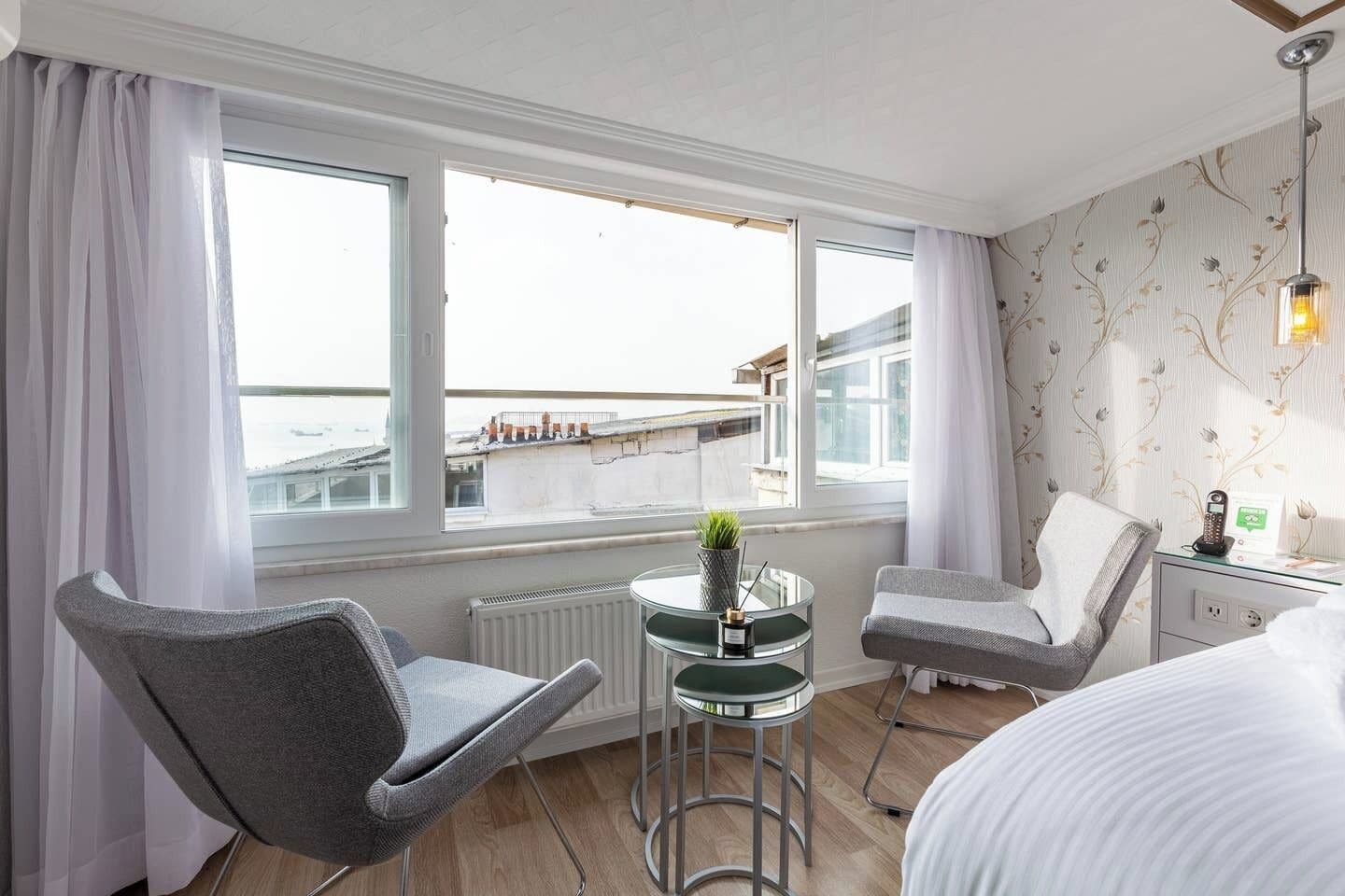 Quarto de hotel com cama, duas cadeiras, uma mesinha com uma planta e vista para a paisagem urbana através de grandes janelas.