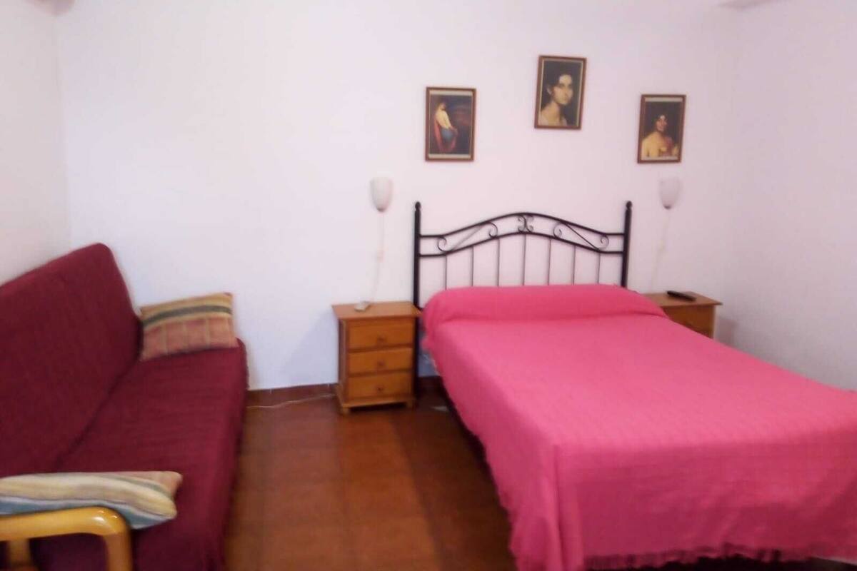 A bedroom with a single bed, a wooden nightstand, a red sofa, and a small table with a lamp.