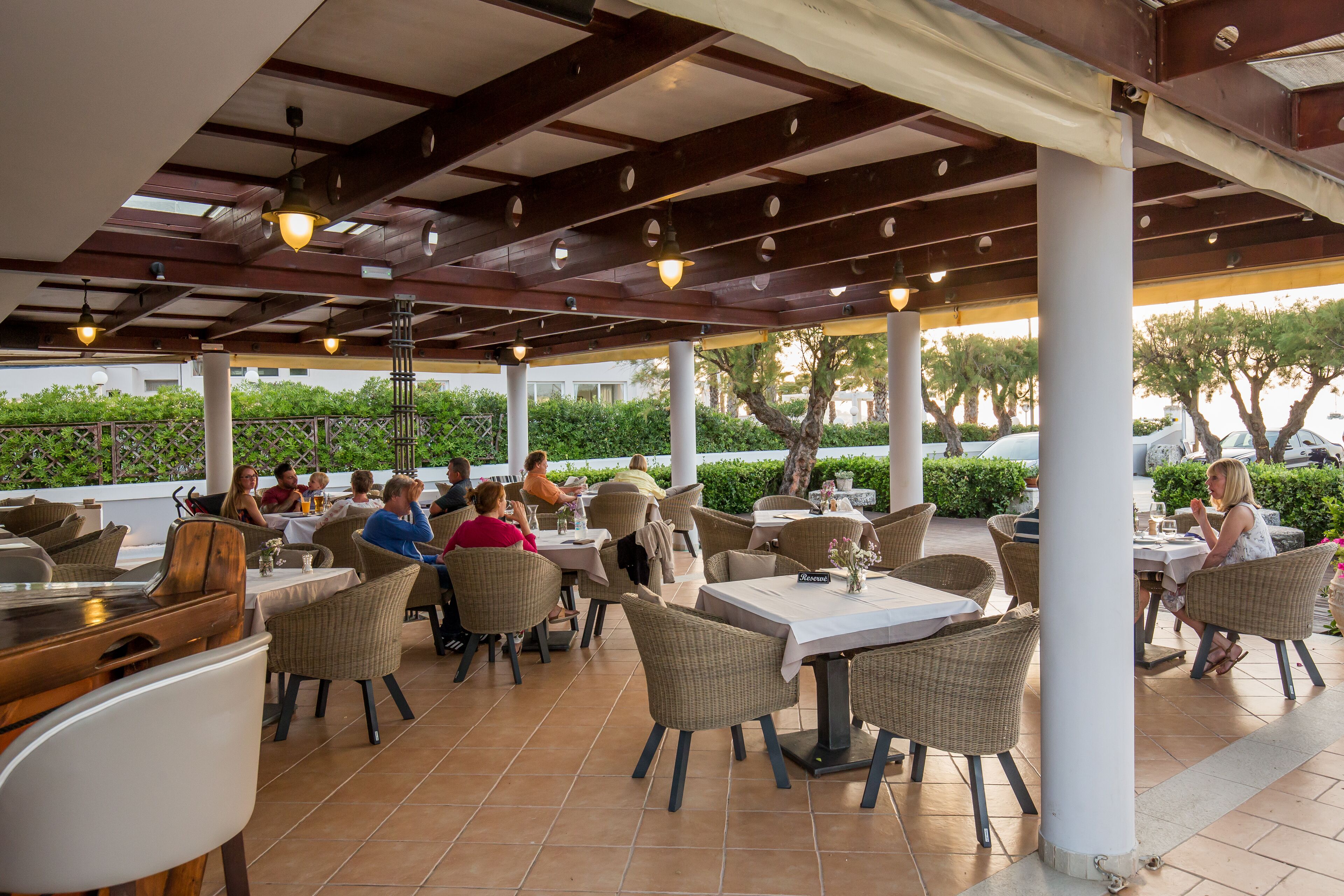 Un área de comedor al aire libre con sillas y mesas de mimbre, una barra de madera y un techo cubierto con luces colgantes.