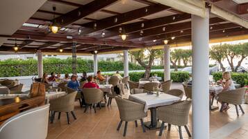 Un área de comedor al aire libre con sillas y mesas de mimbre, una barra de madera y un techo cubierto con luces colgantes.
