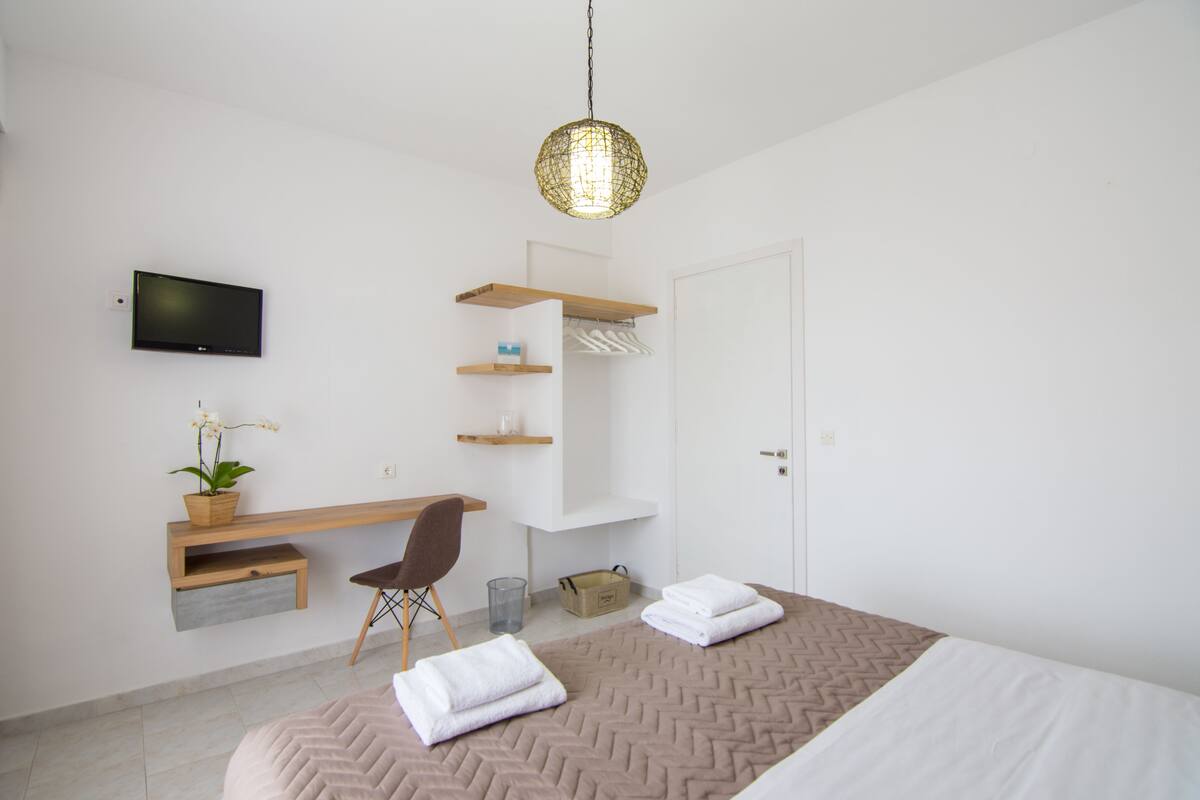 A modern bedroom with a bed, a desk, a chair, a TV, and a hanging light fixture.
