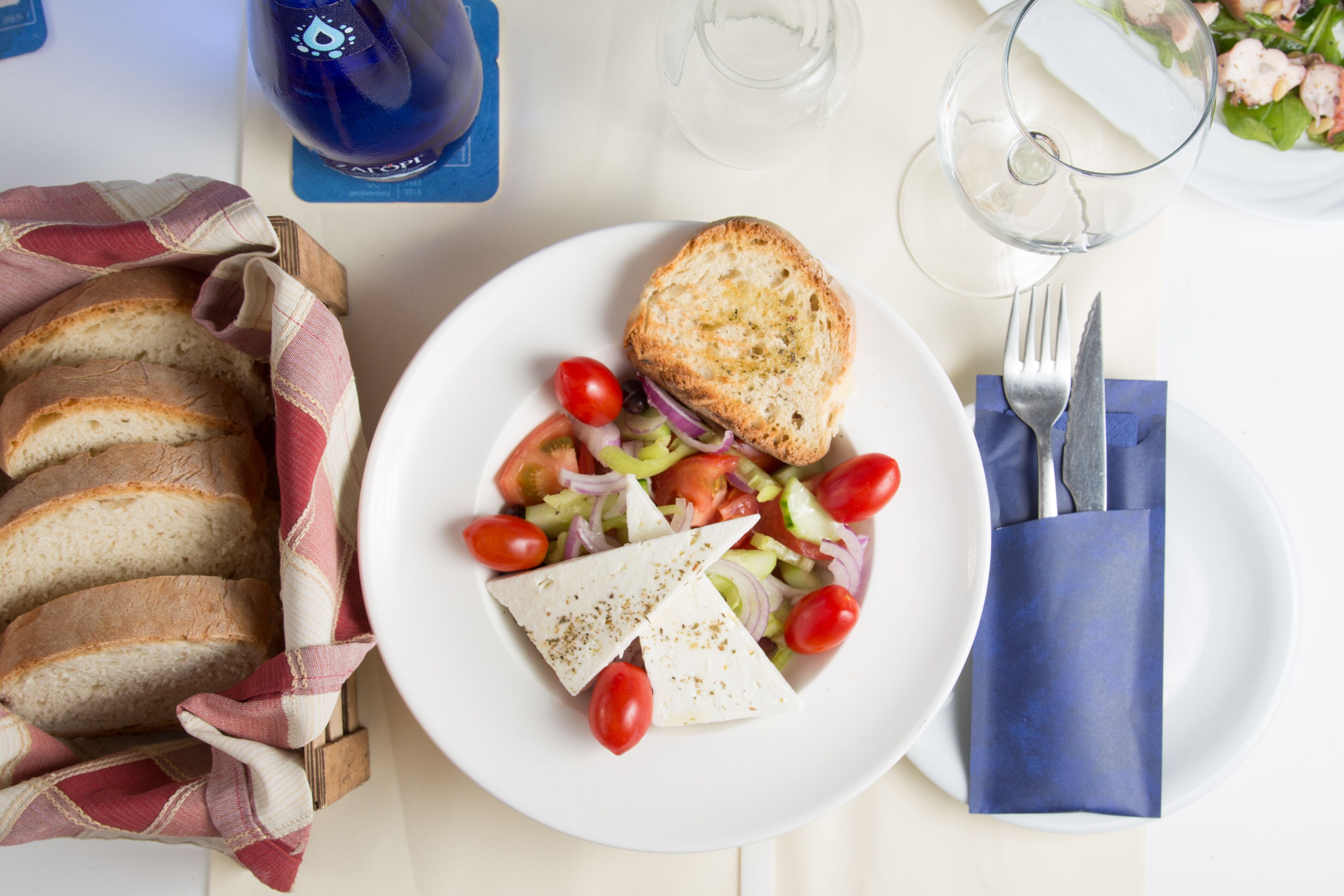 Un plato con ensalada, queso y pan, un vaso de agua y una servilleta con cubiertos sobre una mesa blanca.