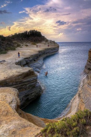 A coastal landscape with a rocky shoreline, a narrow water channel, and people walking along the cliffs.