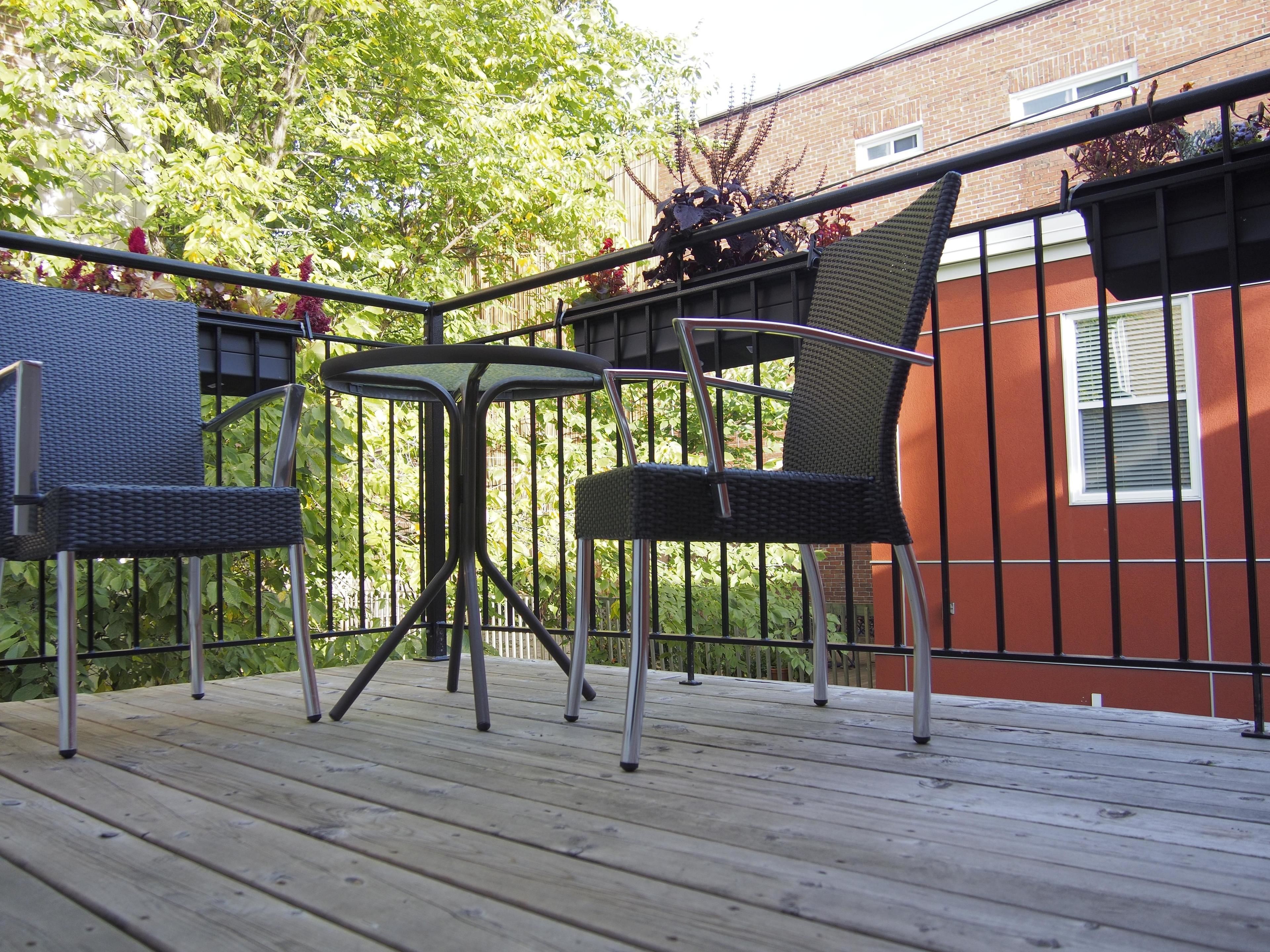A deck with two wicker chairs and a small table, surrounded by a metal railing and a brick wall.