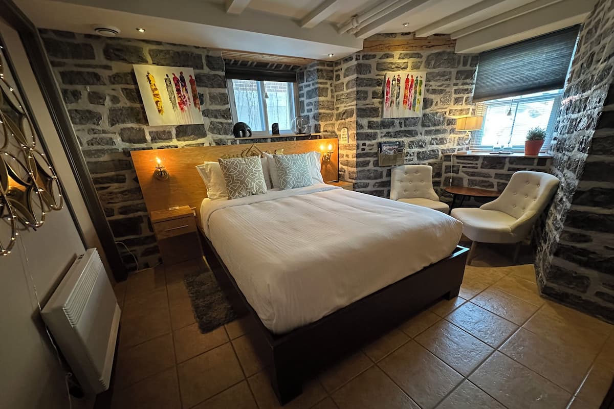 A bedroom with a stone wall, a bed with patterned pillows, a chair, and a small table.