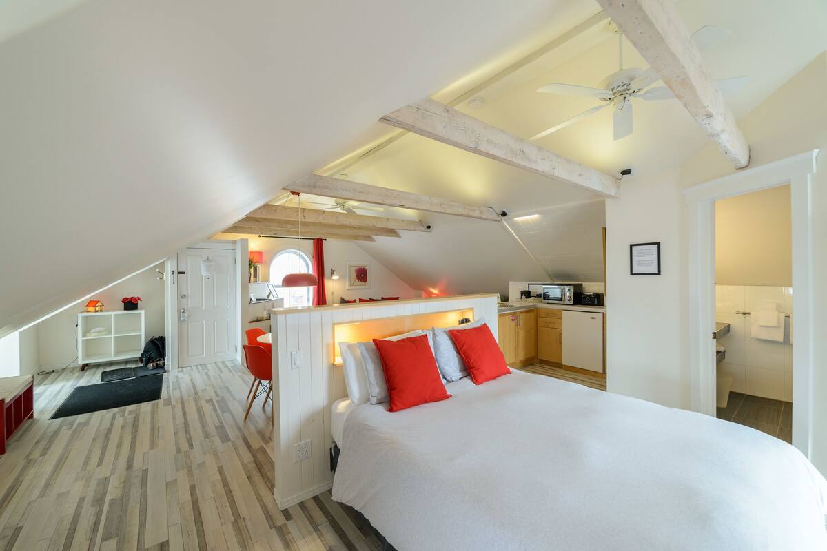 A bedroom with a large bed, a ceiling fan, and a kitchenette in the background.