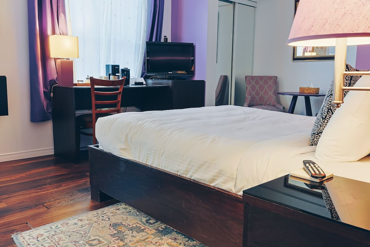 A hotel room with a bed, a desk with a television, a chair, a small table, and a lamp.