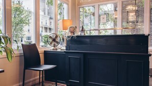 Une pièce avec un piano, une chaise, un ventilateur et une vue sur les immeubles à travers de grandes fenêtres.