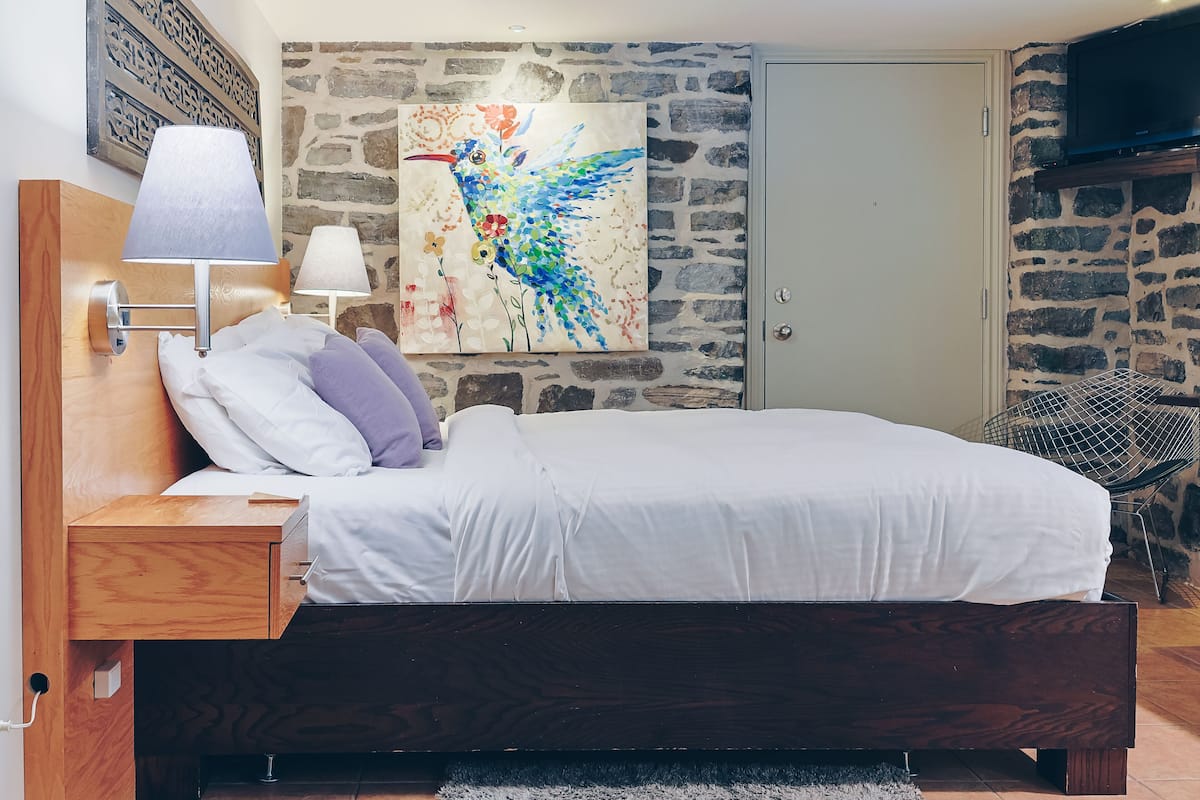 A bedroom with a wooden headboard, a bed with white bedding, a nightstand with a lamp, a stone wall, and a painting of a bird.
