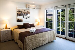 Un dormitorio con una cama grande, dos mesitas de noche, un aire acondicionado instalado en la pared y un ventanal corredero que da a un balcón con vegetación.
